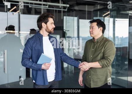 Zwei verschiedene Geschäftsleute schütteln die Hand und schließen eine Partnerschaftsvereinbarung in einem modernen Glasbüro, die erfolgreiche Verhandlungen, Zusammenarbeit und multikulturelle Teamarbeit repräsentieren Stockfoto