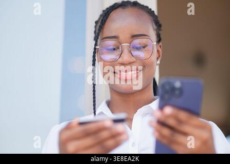 Lächelnde junge Schwarze Frau in Brille mit Kreditkarte und Smartphone für Online-Zahlung. Mobiles Banking, digitales Shopping, kontaktloses Finanzwesen, umweltfreundlich Stockfoto
