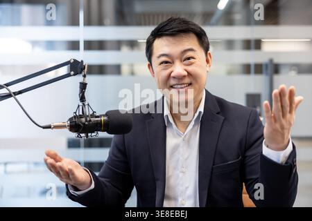 Asiatischer Geschäftsmann, der selbstbewusst Gesten in ein professionelles Studio-Mikrofon einnimmt und ansprechende Audioinhalte für einen Podcast oder ein Online-Interview erstellt Stockfoto