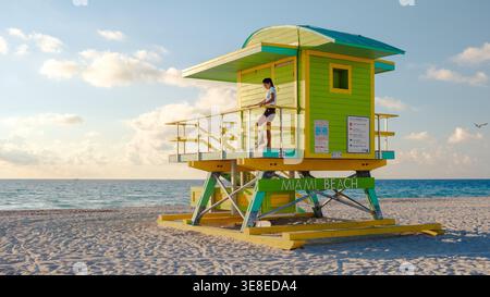 Ein farbenfroher Rettungsschwimmer-Turm bewacht Miami Beach, während die Sonne aufgeht und warmes Licht auf das Sandstrand wirft. Eine Einzelfigur genießt die ruhige morgendliche Atmosphäre. Stockfoto