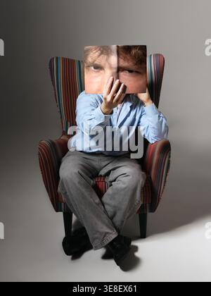 Junger Mann, der im Sessel sitzt und ein Notizbuch mit bedrucktem Studio-Porträt hält. Stockfoto