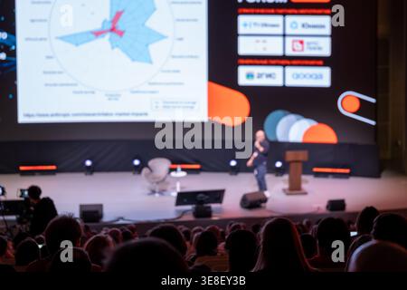 Konferenzpräsentation auf der Bühne mit Sprecher, Grafik auf Großbildleinwand, aufmerksames Publikum in dimmem Auditorium. Stockfoto