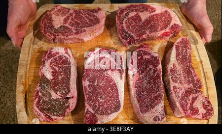 Rohe Ribeye-Steaks, gewürzt auf Holzbrett. Nahaufnahme mehrerer roher marmorierter Ribeye-Steaks, die mit Salz und Pfeffer auf einem hölzernen Schneidebrett gewürzt wurden Stockfoto