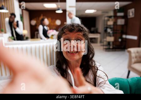 Lächelnde ältere Frau in der Hotellounge macht Selfie auf ihrem Handy, während das Personal anderen Gästen im Hintergrund hilft. Pensionierte Seniorin, die lustige Ferien auf dem Handy fängt. Stockfoto