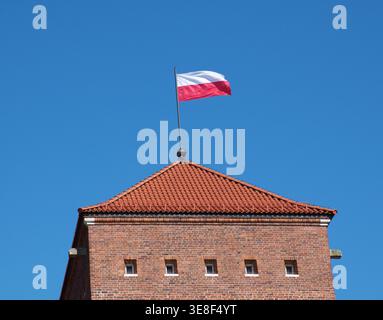 Flacher Blick auf die polnische Nationalflagge, die auf einem historischen Backsteinturm mit rotem Ziegeldach vor einem klaren blauen Himmel winkt. Minimalistisches Konzept. Stockfoto