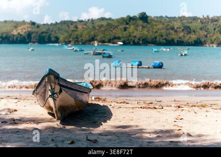 Ein farbenfrohes lokales Fischerboot, das auf dem Sand im Vordergrund liegt, mit einer Bucht voller verankerter Boote und üppiger Hügel im Hintergrund. Stockfoto