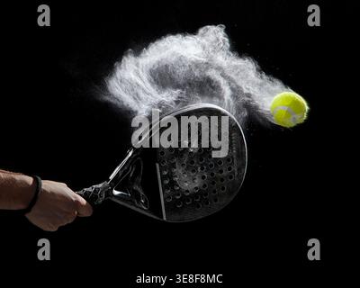 Ein Padel-Schläger trifft einen Tennisball in der Luft und erzeugt eine Staubwolke, die in einem spektakulären Schnappschuss vor schwarzem Hintergrund erfasst wird. Stockfoto