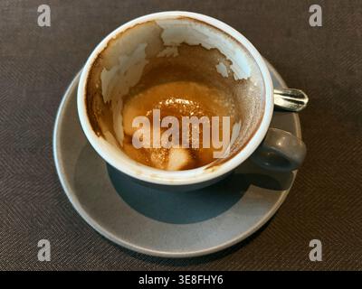 Nahaufnahme einer leeren Kaffeetasse mit Schaumrückständen auf der Untertasse in einer Café-Umgebung. Konzept von Konsum, Routine und Café-Lebensstil. Stockfoto