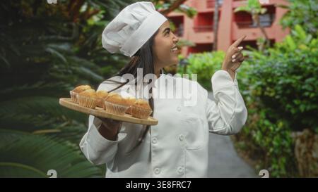 Die Chefin in weißem Mantel hält mit einer Hand ein hölzernes Muffins-Tablett und zeigt mit dem Finger auf den Weg zum Bau des Hofes; Backfreude. Stockfoto