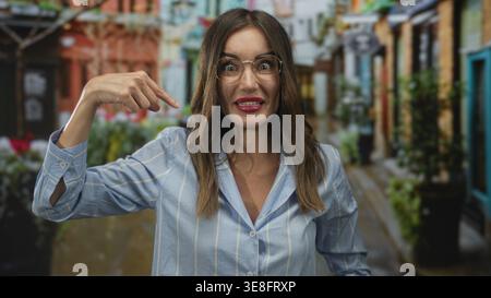 Junge hispanische Frau mit Brille zeigt beide Zeigefinger auf sich, während sie auf der belebten Straße lächelt; Selbstvertrauen. Stockfoto