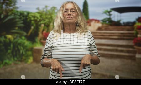 Eine ältere Frau zeigt mit den Zeigefingern auf eine Steintreppe im Gebäude, trägt ein gestreiftes Hemd und eine Brille; trotzt dem Stoizismus. Stockfoto
