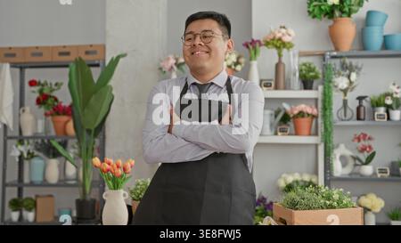 Blumenhändler mit Schürze und Krawatte, der einen orangen Tulpenstrauß hält, Arme überkreuzt und lächelt in einem Gebäude mit Vasen- und Topfpflanzen-Regalen; Stockfoto