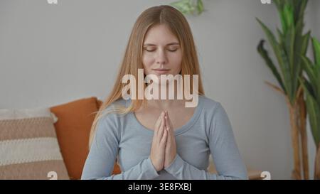 Junge Frau mit zusammengedrückten Händen in Gebetsgeste auf Sofa im Studio, Augen geschlossen und entspannt, Zimmerpflanze und Kissen sichtbar; Gelassenheit Mindfu Stockfoto