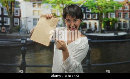 Die Frau zeigt mit dem Finger auf die Papiertüte auf der Kanalstraße, hält sie hoch und lächelt breit; Überraschung. Stockfoto