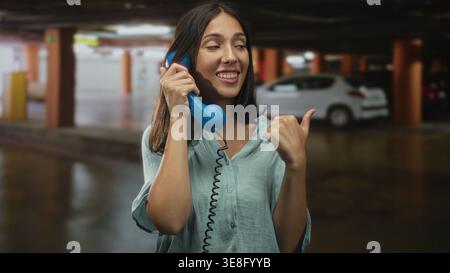 Junge brünette Frau, die einen blauen Telefonhörer hält, zeigt den Daumen über die Schulter in einem dunklen Parkhaus in der Nähe geparkter Autos; verspielter Unfug. Stockfoto
