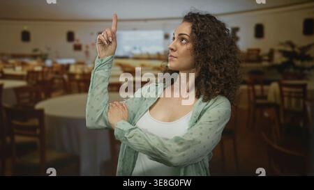 Frau zeigt den Zeigefinger im Speisesaal des Restaurants in der Nähe runder Tische mit umgeklappten Armen und neutralem Blick nach oben; Selbstbewusstsein. Stockfoto