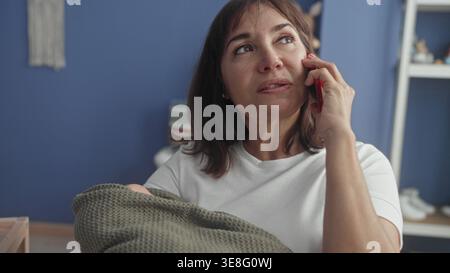 Frau, die das Smartphone am Ohr hält, während sie eine eingewickelte Babydecke neben einer hölzernen Krippe im Kinderzimmer hält; müdes Mitgefühl. Stockfoto