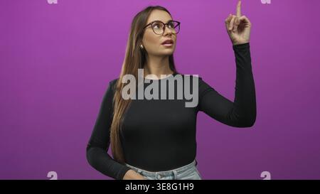 Die junge Frau zeigt beide Zeigefinger nach oben, trägt eine Brille und zeigt Finger und Gesicht in einem Studio; Selbstvertrauen feiern. Stockfoto