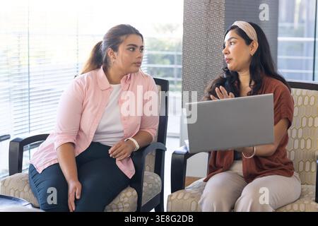 Verschiedene weibliche Kollegen diskutieren das Projekt in der Bürolounge, während sie in Sesseln mit Laptop sitzen Stockfoto