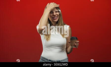 Frau in weißem Tanktop und Jeans, die eine Brille trägt, die eine Wellpappkaffeetasse mit Hand auf der Stirn im Studio hält; peinliches Lächeln. Stockfoto