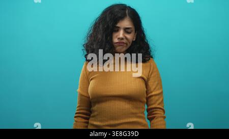 Junge arabische Frau Gesicht mit geschlossenen Augen und schmerzhaftem Ausdruck in Senfpullover in türkisfarbenem Studio, leicht geneigter Kopf und nach unten gerichtetem Blick; Traurigkeit Stockfoto