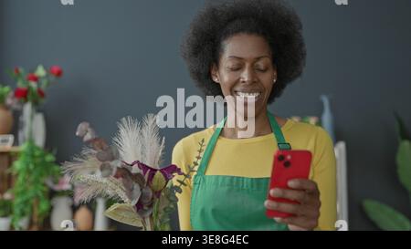 Floristin mit grüner Schürze hält Smartphone und Strauß aus getrockneten Gräsern und Eukalyptus im Blumengeschäft-Studio; Freude an kleinen Unternehmen. Stockfoto