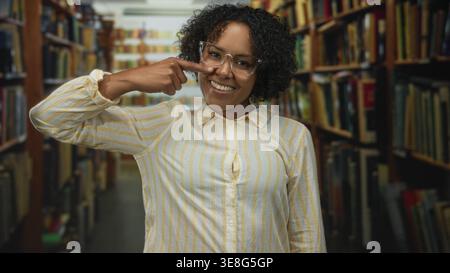 Frau mit lockigem Haar und Brille zeigt mit dem Finger auf Brille im alten Bibliotheksgebäude; Selbstvertrauen fokussiert. Stockfoto