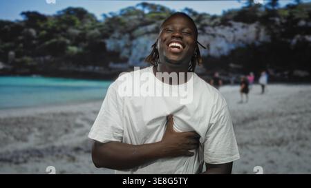 Mann lacht mit Hand auf Brust an einem Sandstrand neben türkisfarbenem Meer und felsigen Klippen, trägt weißes T-Shirt und Dreadlocks in der Strandszene; Stockfoto