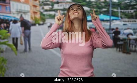 Frau zeigt beide Zeigefinger nach oben und zeigt Finger und Handflächen auf einer belebten Straßenpromenade; hoffe Neugier. Stockfoto
