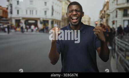 Mann mit geballten Fäusten, schreiend und lachend auf einer belebten europäischen Straße, mit Brille und Marinepolo, mit aufgerissenem Mund in einer kurzen Sequenz; Stockfoto