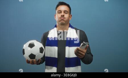 Mann, der Fußball und Smartphone hält, die Lippen und die Augen mit einem blauen und weißen Schal im Studio verschlossen hat; Freude an der Begeisterung der Fans. Stockfoto
