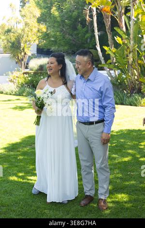 Ein Paar älterer asiatischer Mann und eine Braut, die Hochzeitskleid und Schleier tragen, lächelnd im Garten mit Blumenstrauß Stockfoto