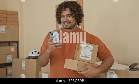 Mann in orangefarbenem Hemd hält Kartons und Smartphone, lächelt, während er in einem Pappraum des Gebäudes telefoniert; Optimismus für kleine Unternehmen. Stockfoto