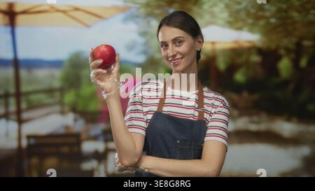 Frau hält Apfel mit Handschuhen auf der Terrasse des Restaurants, trägt Schürze und gestreiftes Hemd; Selbstvertrauen gesunde Ernährung. Stockfoto