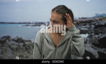 Junge hispanische Frau, die mit der Hand das Ohr schröpft und im Studio am Meer schielt, mit grauem Kapuzenpullover; frustrierte Hörschwierigkeiten. Stockfoto
