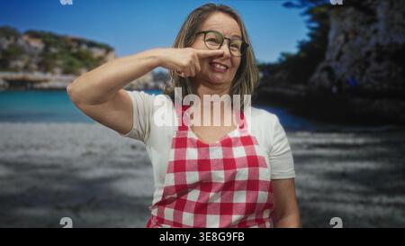 Hispanische Seniorin mit grauen Haaren und Brille, die eine rote karierte Schürze trägt, zeigt den Finger an die Nase und lächelt vor einem Studio-Strand-Hintergrund; fröhlich. Stockfoto