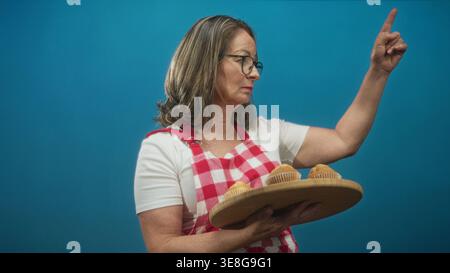 Hispanische Köchin, die Muffins auf einem Holztablett hält, zeigt den Zeigefinger nach oben im Studio; hausgemachter Stolz. Stockfoto