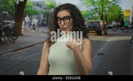 Frau zeigt auf sonniger Straße mit lockigem Haar und Brille den Finger auf sich; verweigert die Ablehnung. Stockfoto