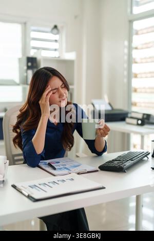Überforderte Erwachsene asiatische Mitarbeiterin, die unter intensivem Kopfschmerz am Arbeitsplatz leidet. Sie hält eine Kaffeetasse am Schreibtisch mit Business-Charts. Stockfoto