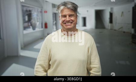 Mann mit lächelndem Gesicht in cremefarbenem Pullover, mit entspannten Händen nach vorne gerichtet in einem hellen, leeren Studio mit Treppenhaus und großen Fenstern Stockfoto