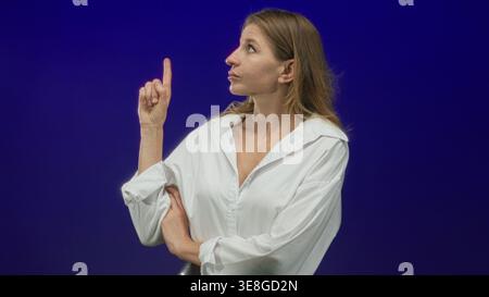 Die Frau zeigt den Finger im blauen Studio nach oben, trägt ein weißes Hemd mit gerollten Ärmeln und gefalteten Armen, blickt mit konzentriertem Ausdruck nach oben; Neugier. Stockfoto
