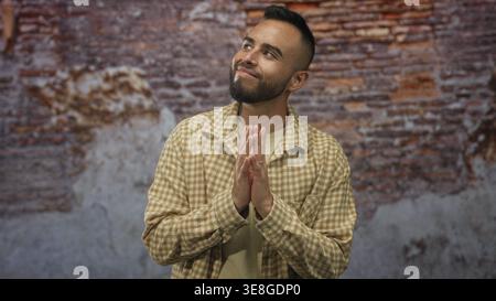 Junger hispanischer Mann mit gefesselten Händen und leichtem Lächeln, der aufblickte, kariertes Flanellhemd mit Ringen trug und vor einer strukturierten Ziegelmauer in Bu stand Stockfoto