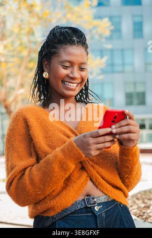 Eine entzückte junge Frau, die mit ihrem Smartphone interagiert und hell lächelt, während sie ihre Zeit im Freien mit Freunden und Familie genießt und Momente festnimmt Stockfoto