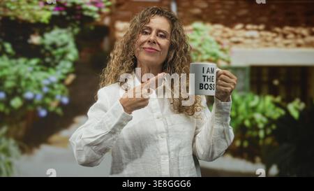 Die hispanische Frau im mittleren Alter zeigt mit dem Finger auf den Becher und liest: ich bin der Boss in einem Gebäude, lächelt und hält den Becher in der rechten Hand; Selbstvertrauen stärken Stockfoto