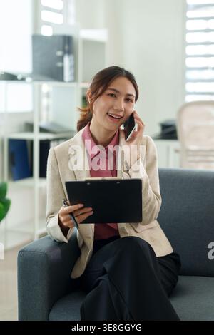 Erwachsener asiatischer Mitarbeiter kommuniziert über Smartphone verwaltet Aufgaben Halten des Dokumentenstifts Lächeln am Arbeitsplatz Einstellung der Fernverbindung Stockfoto