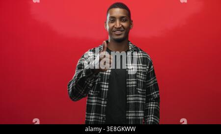 Junger Mann mit schwarzem kariertem Hemd, der Daumen nach oben zeigt und mit der Hand in einem roten Studio zeigt; Zustimmungsermutigung. Stockfoto