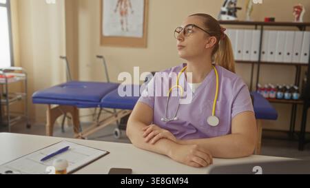 Frau Ärztin in lila Peelings mit gelbem Stethoskop am Schreibtisch, Hände gefaltet neben Klemmbrett und Untersuchungstisch im Gebäude; nachdenkliche Pflege. Stockfoto