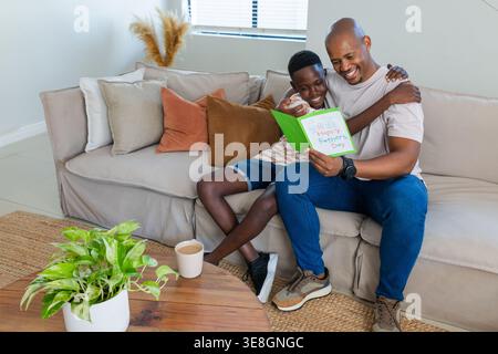 Vater und männliches Kind sitzen im Wohnzimmer auf dem Sofa und halten die grüne Happy Vatertag-Karte Stockfoto
