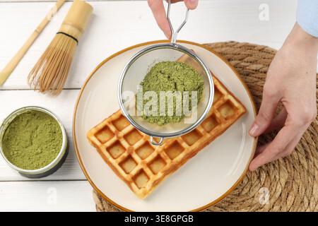 Frau siebt Matcha-Pulver auf Waffeln an weißem Holztisch, Blick von oben Stockfoto