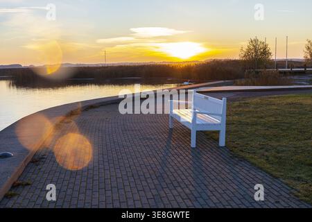 Leere Bank an einer Strandpromenade bei Sonnenaufgang in Haapsalu, Estland. Ruhige Küstenlandschaft mit warmem Morgenlicht und friedlicher Atmosphäre. Stockfoto